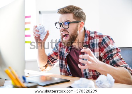 Angry crazy modern designer in glasses with beard yelling and crumpling paper on his workplace Royalty-Free Stock Photo #370320299