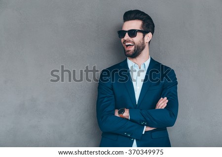 Stylish smile. Cheerful young handsome man in sunglasses keeping arms crossed and looking away with smile while standing against grey background Royalty-Free Stock Photo #370349975