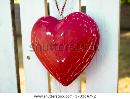 Closeup of weathered red heart hanging from white rustic wooden fence.