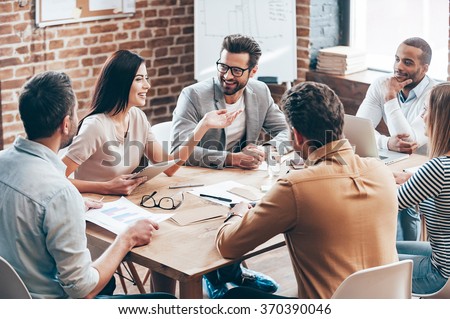 Making great decisions. Young beautiful woman gesturing and discussing something with smile while her coworkers listening to her sitting at the office table Royalty-Free Stock Photo #370390046
