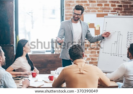 Take a look at our results! Handsome young man in glasses standing near whiteboard and pointing on the chart while his coworkers listening and sitting at the table 