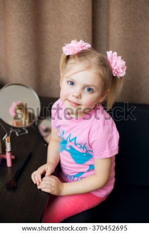 little cute blonde girl smiling three - four years in a pink dress with two tails sits near the mirror with a set of cosmetics and doing makeup, lipstick, cheek colors large fluffy brush for powder