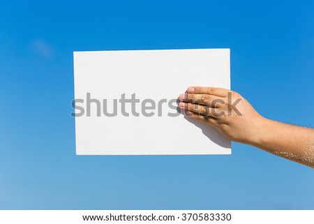 Blank white board in hand against blue sky background. Tropical summer vacation concept
