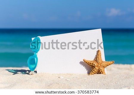 Blank white board, swimming glasses and starfish on sand against turquoise caribbean sea water. Tropical summer vacation concept
