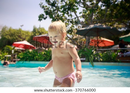 closeup small blonde girl stands and holds metal pole in shallow water of hotel pool against colourful parasols