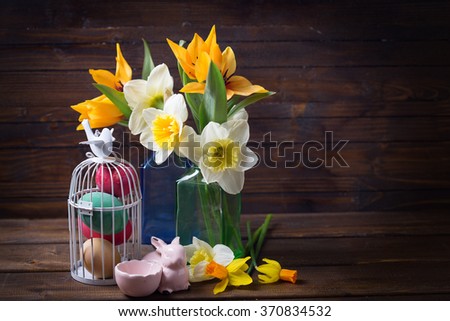 Colorful easter eggs in bird cage, fresh tulips and narcissus in vases on on aged dark wooden background. Easter background. Place for text.
