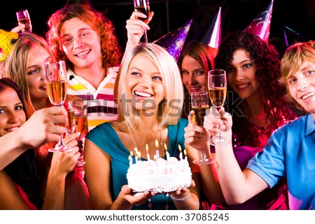 Portrait of joyful girl holding birthday cake surrounded by friends with flutes of champagne