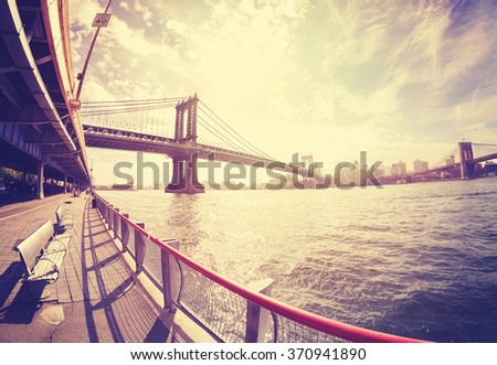 Vintage stylized fisheye lens photo of bench at Hudson River bank and Manhattan Bridge in distance against sun, NYC, USA.