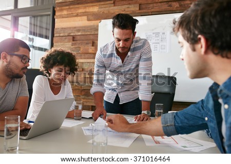 Young man discussing market research with colleagues in a meeting. Team of young professionals having a meeting in conference room looking at documents. Royalty-Free Stock Photo #371016464