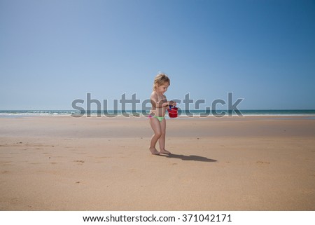 two years old back blonde baby with pink and green swimsuit walking with red plastic bucket in hand at golden sand beach seaside in Cadiz Andalusia Spain