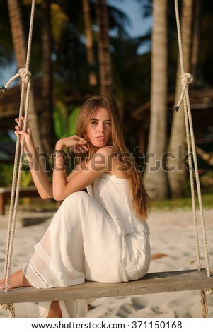 Beautiful model girl with long dark hair in long white dress sits on the sandy beach with green exotic palm trees in sunglasess and dreams with a smile