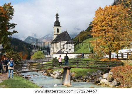 People enjoying the beautiful scenery on a bridge in front of a church with foggy mountains in the background ~ Magnificent autumn scenery of Bavarian countryside in Ramsau Germany