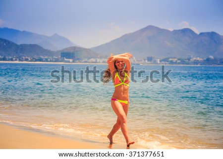 blonde slim female gymnast in bikini poses crossing arms over head on sea edge of beach smiles against resort city