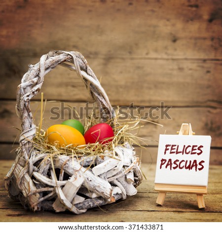 white basket with easter eggs, canvas with spanish text felices pascuas, which means happy easter, wooden background
