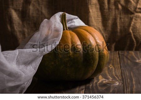 Ripe round oblate orange pumpkin with smooth segmented surface and green formless blotch with white gauze on wooden table on burlap background, horizontal photo