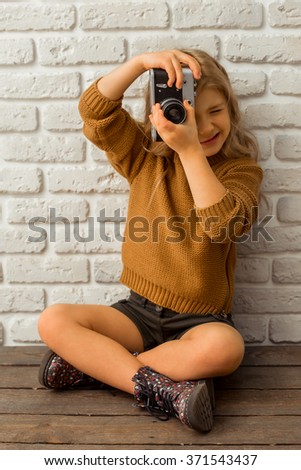 Pretty little blonde girl making a photo while sitting cross-legged against white brick wall