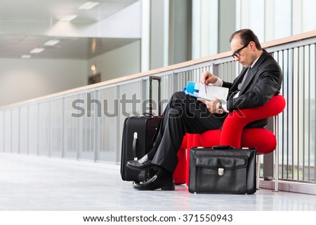 Business man with trolley and suitcase reading business catalog on red armchair at exhibition lobby