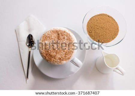 Cup of hot coffee in white ceramic cup and plate with a small jar of syrup and sugar on a white table.