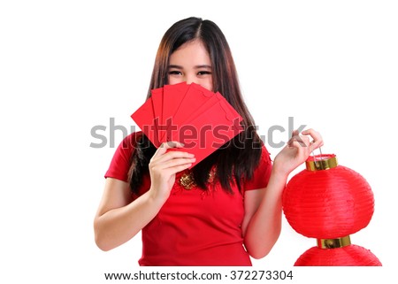 Cute shy Asian teenager in cheongsam dress covering her face with red envelopes while holding a Chinese lantern, isolated on white background