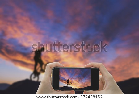Taking photo on smart phone concept.Man riding a bike performing a trick against sunset sky. 