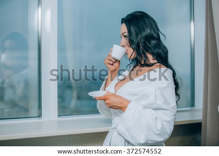 Getting warm with fresh coffee. Beautiful young woman in white bathrobe drinking coffee and looking through a window