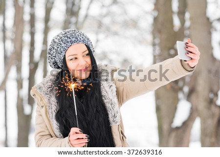 Closeup portrait of cute young woman in beanie taking a selfie on smartphone, outdoors in winter, holding a sparkler. Happy young woman photographing herself using cellphone outdoors in wintertime.