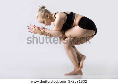 Sporty beautiful young woman practicing yoga, doing Eagle Fold Pose, variation of Garudasana, exercise for ankles, calves, thighs, hips and shoulders, working out wearing black sportswear, studio