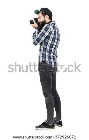 Side view of hipster in plaid shirt and baseball cap taking photo with dslr camera. Full body length portrait isolated over white studio background.