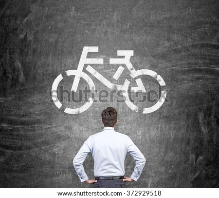 man with hands on hips standing in front of a picture of a bicycle on the blackboard. Back view. Concept of eco traveling. Royalty-Free Stock Photo #372929518