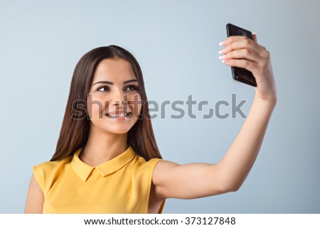 Photo of beautiful young business woman standing near gray background. Woman with yellow shirt making selfie with mobile phone and smiling