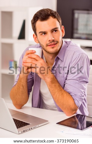 Young attractive businessman in casual clothes drinking coffee and dreaming while working in office