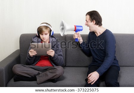 Tween son in headphones looks at the digital tablet display while his father yells at him through a megaphone Royalty-Free Stock Photo #373225075