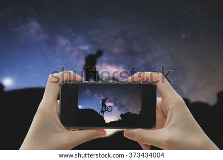 Taking photo on smart phone concept.Man riding a bike performing a trick against on Mountain with Milky way. 