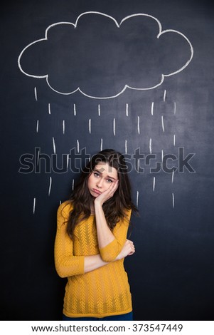 Sad cute young woman standing under raincloud and rain drawn above her on a blackboard background