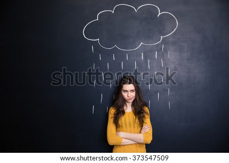 Unpleased young woman with raincloud drawn over her on a blackboard background standing with arms crossed  Royalty-Free Stock Photo #373547509