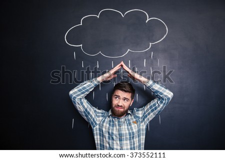 Frowning irritated young man holding hands above head and covering from drawn rain from raincloud over blackboard background Royalty-Free Stock Photo #373552111