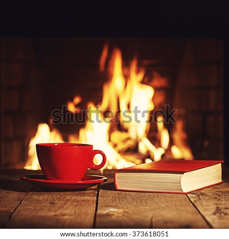 Red cup of coffee or tea and old book on wooden table near  fireplace. Winter and Christmas holiday concept. Photo with retro filter effect. Royalty-Free Stock Photo #373618051