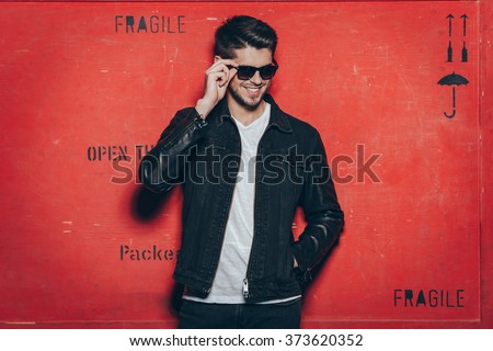 Cheerful and trendy look. Handsome young man adjusting his sunglasses and smiling while standing against red background