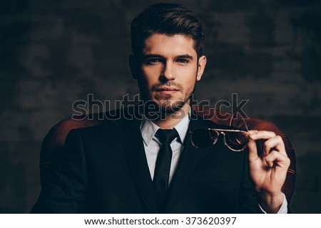Breathtaking look.Portrait of young handsome man in suit holding his sunglasses and looking at camera while sitting in leather chair against dark grey background Royalty-Free Stock Photo #373620397