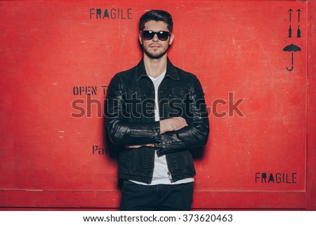 Trendy handsome. Handsome young man in sunglasses keeping arms crossed and looking at camera while standing against red background