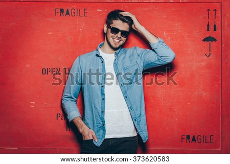 Cheerful and trendy.Handsome young cheerful man in sunglasses keeping hand in hair and looking at camera with smile while standing against red background