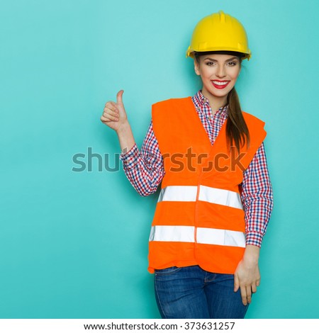 Smiling young woman in orange reflective vest, jeans and yellow hardhat showing thumb up and looking at camera. Three quarter length studio shot on turquoise background.
