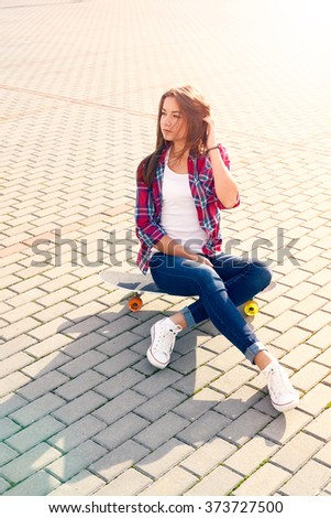 Young girl with long hair in checkered shirt, blue jeans, white sneakers sitting on skateboard, looking away. Youth lifestyle.Urban landscape.Sunset, sunny day.