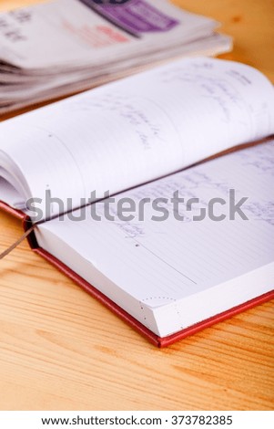 Vertical photo of opened diary with hand writing which is placed on light wooden board front of several newspapers which in background. 