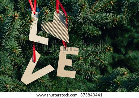 Cardboard LOVE letters hanging on red stripes on green pine tree branches. Holiday postcard template. Space for text, copy, lettering.