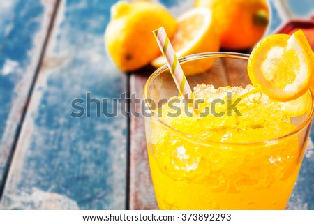 Close Up of Fresh Orange Frozen Granita Slush Drink Garnished with Orange Wedge and Served in Glass with Striped Straw on Weathered Blue Wooden Picnic Table with Oranges in Background - Copy Space Royalty-Free Stock Photo #373892293