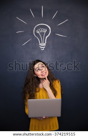Thoughtful smiling young woman holding laptop and looking up over chalkboard background