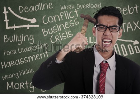 Picture of frustrated businessman hitting his head with hammer in front of blackboard
