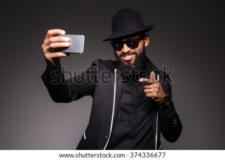 Cheerful afro american man in trendy cloth taking selfie photo on smartphone over black background