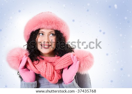 Cheerful woman in pink winter fur cap looking aside with smile on background of snowflakes